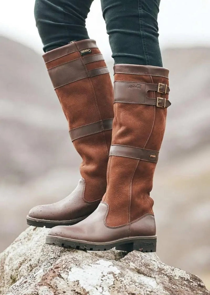 tan and brown leather knee-high waterproof boots with buckle straps on rock